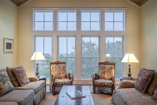 Bright living room with large multi-pane windows, two accent chairs, sofas, and table lamps allowing natural light to fill the space