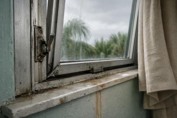 Misaligned aging casement window with fogged glass, loose trim, and water staining inside a Florida bedroom before storm season